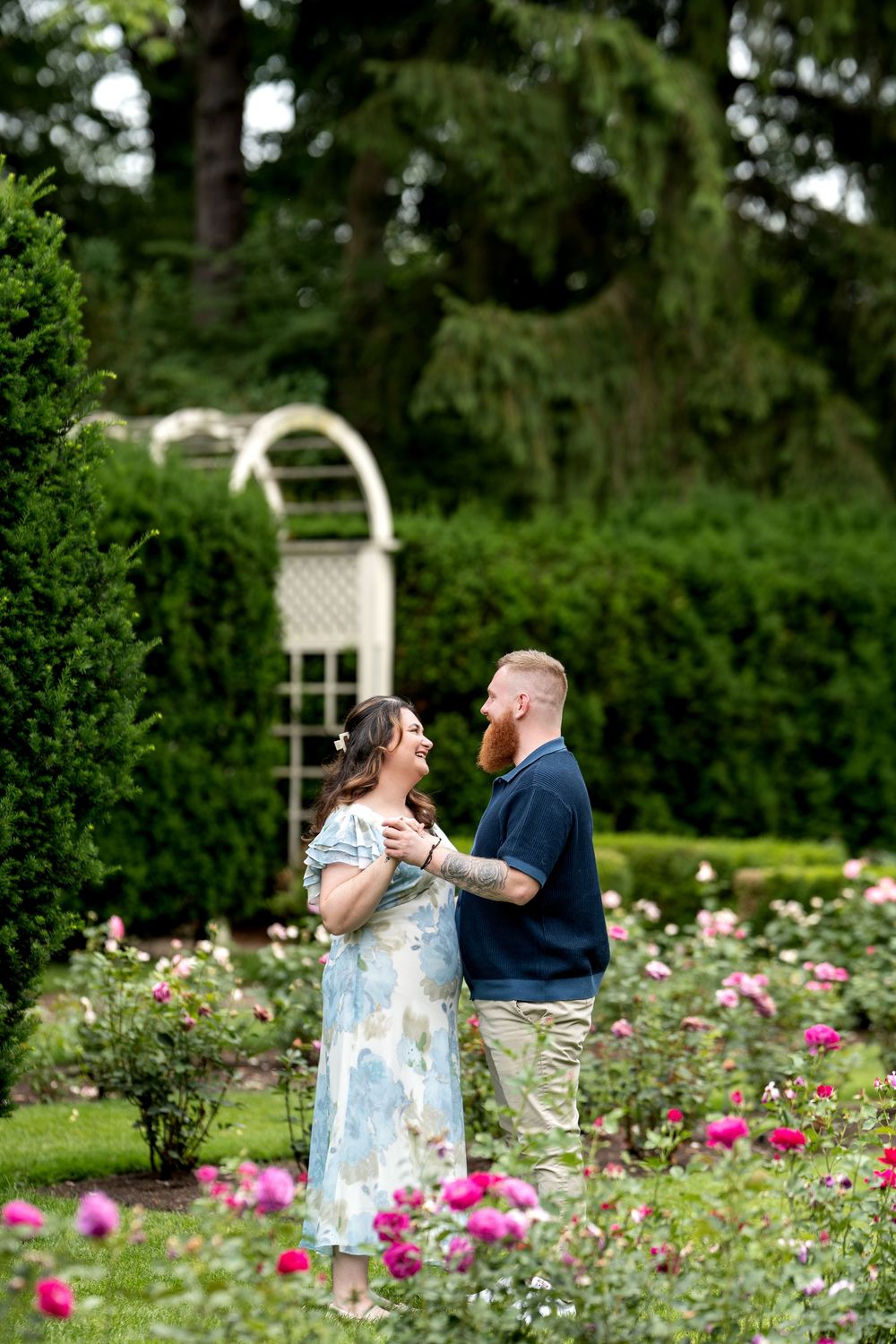 A couple shares a joyful moment in a lush rose garden, the man with a red beard and the woman in a blue floral dress.