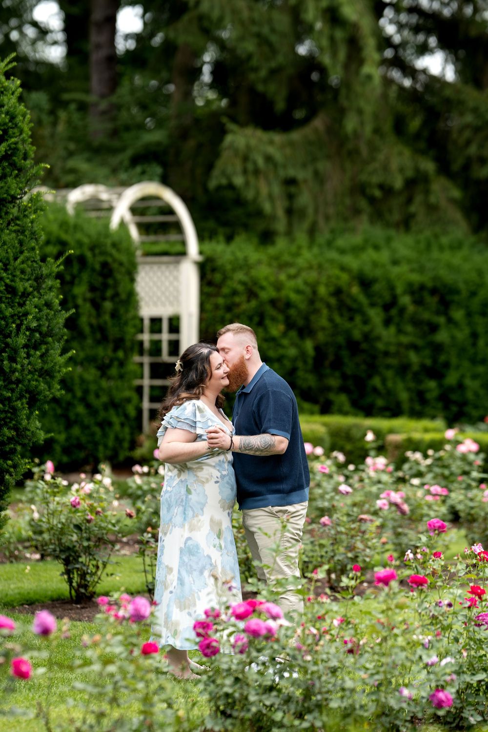 Couple sharing a kiss surrounded by pink roses in a formal garden with hedges and a white trellis.