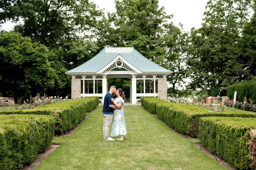 Couple embracing in a formal garden with manicured hedges and a charming stone gazebo in the background.