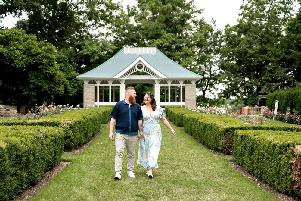 Couple walking hand-in-hand through a lush formal garden with manicured hedges and a white pavilion.