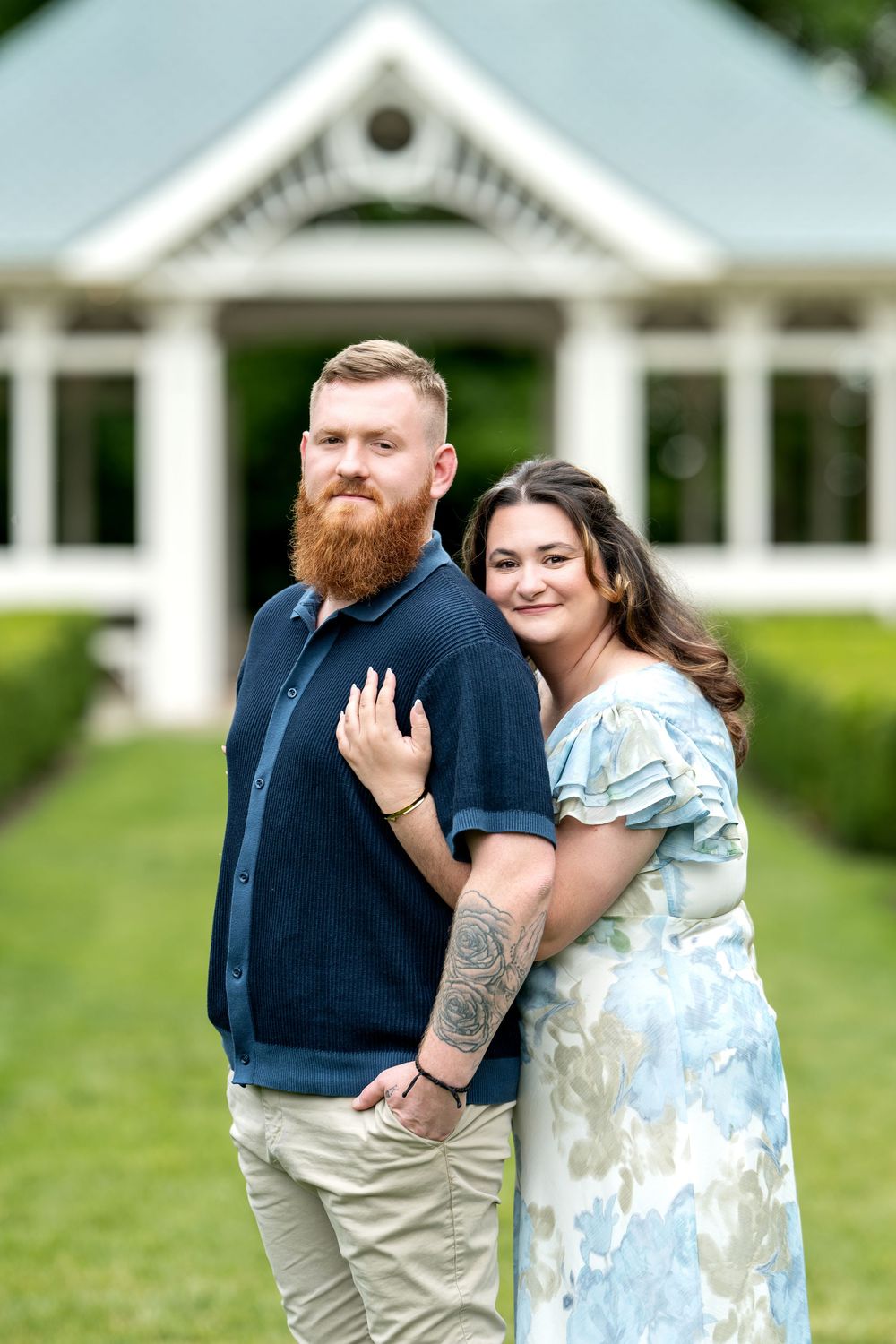A bearded redhead man and brunette woman pose together in front of a white building surrounded by green hedges.