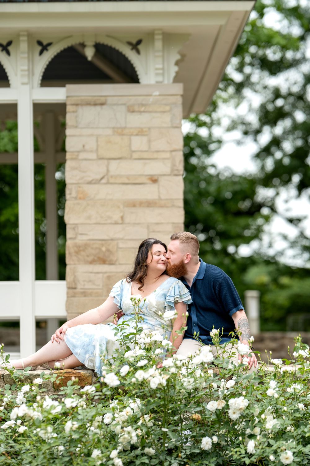 Couple sitting among white roses by a stone gazebo, sharing a kiss in a romantic garden setting.