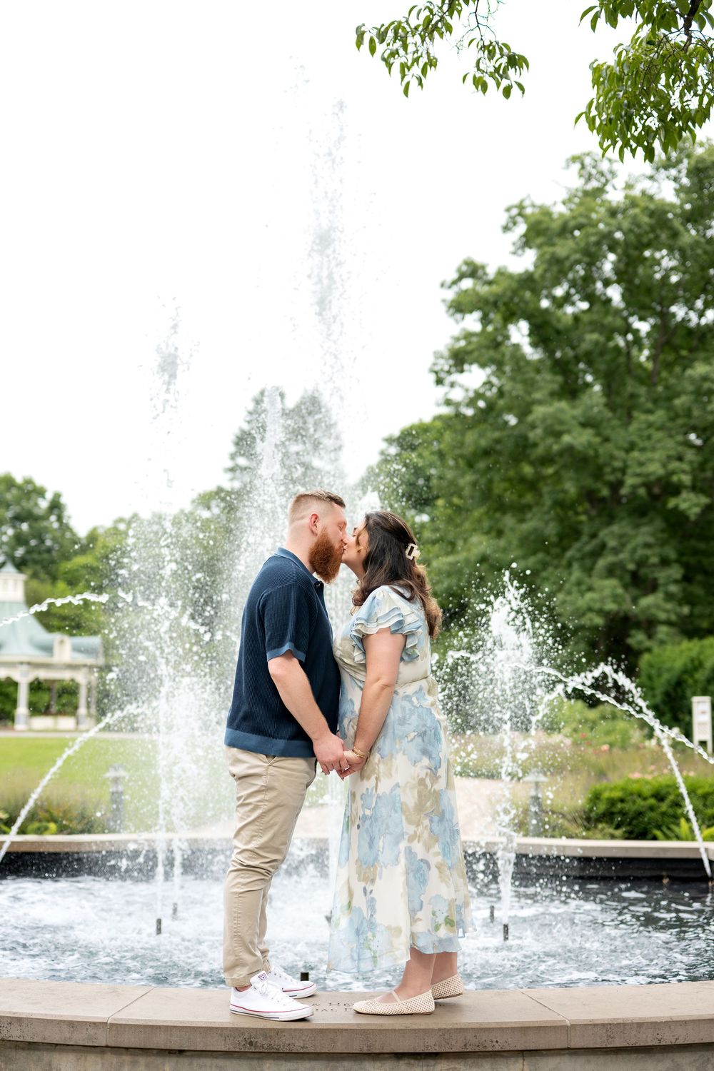 A couple kisses while holding hands in front of a beautiful fountain surrounded by lush green trees.