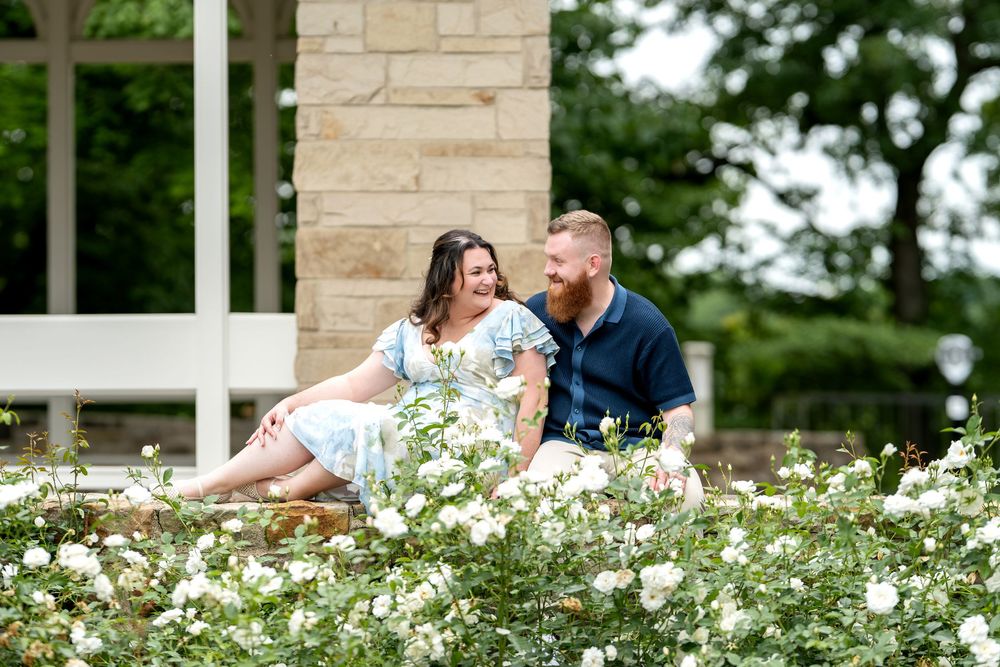 Couple sitting together laughing among white roses in a garden near a stone pillar on a sunny day.