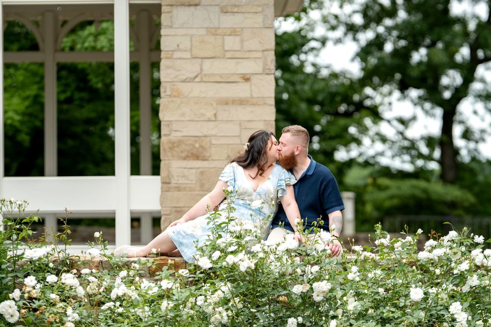 A couple shares a romantic kiss among white roses outside a stone building with lush greenery.