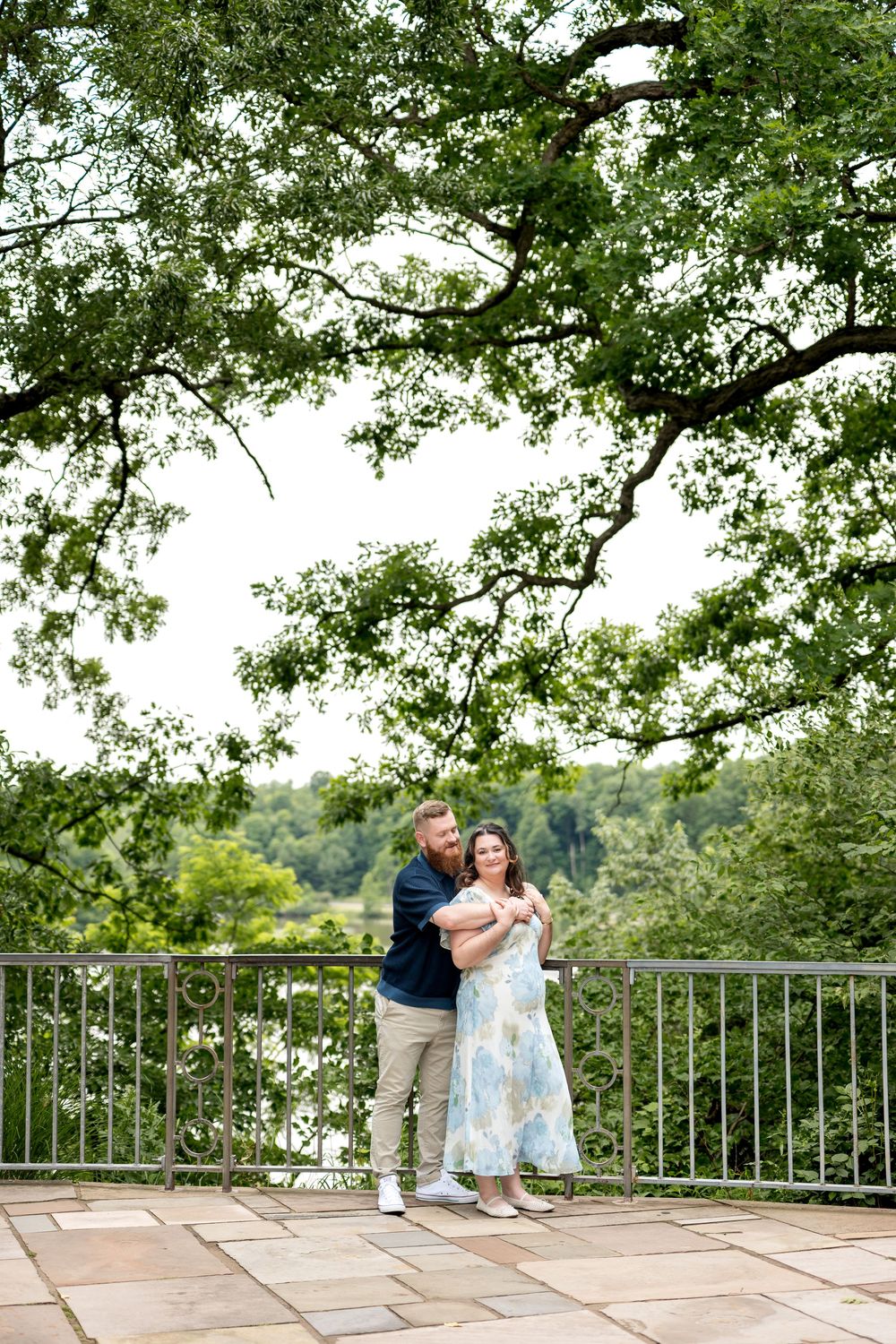 Couple embracing on a stone terrace surrounded by lush green trees and a scenic river view on a cloudy day.