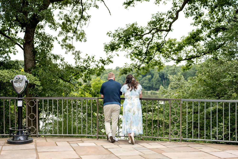 Couple standing at a scenic overlook railing surrounded by lush green trees, enjoying a peaceful outdoor view together.