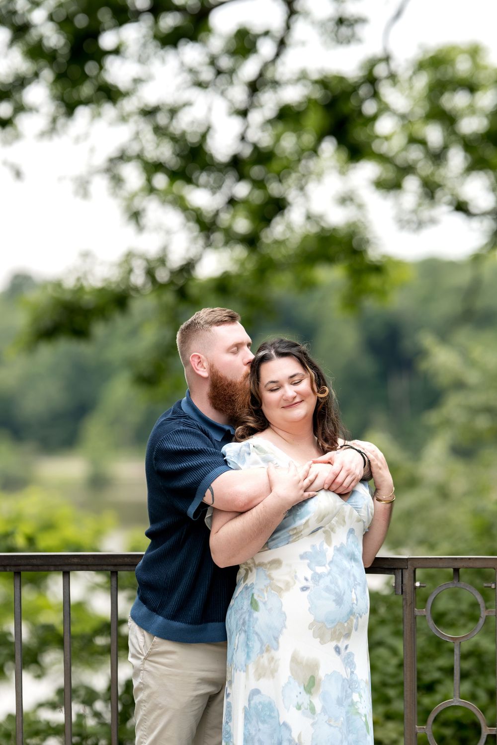 A bearded man in a navy shirt embraces a woman in a floral dress on a balcony, kissing her cheek outdoors.