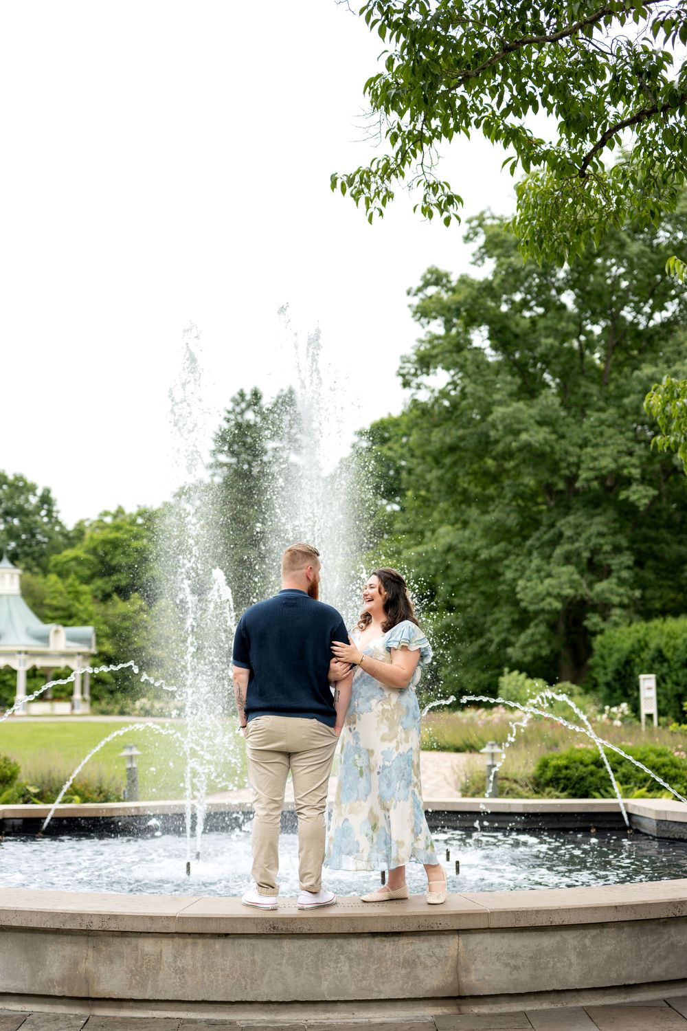 Couple standing by a fountain in a lush garden, woman in floral dress laughing with man in navy shirt.