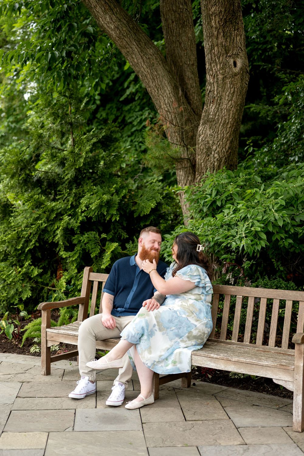 Couple sitting on a wooden bench in a lush garden, sharing a tender moment under a large tree.
