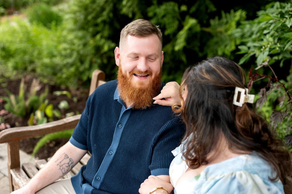A bearded man with tattoos laughs joyfully with a dark-haired woman in a lush garden setting.