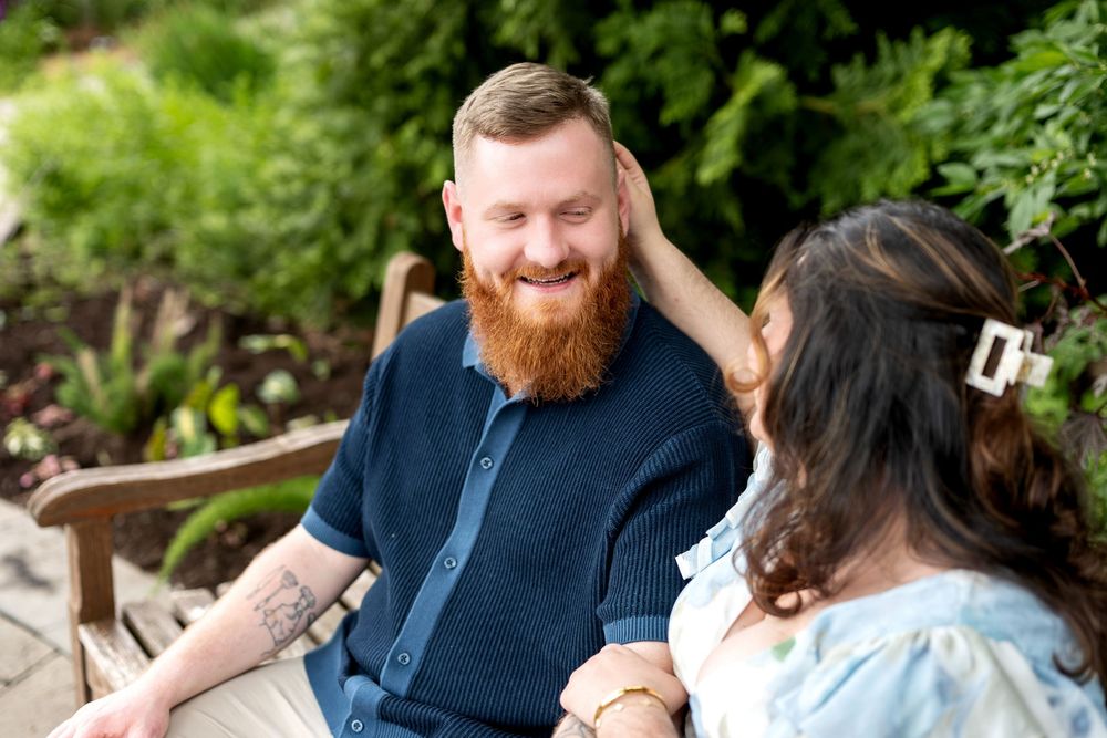 A bearded redhead man smiles warmly at a woman in a garden setting during an outdoor couples engagement photo session.