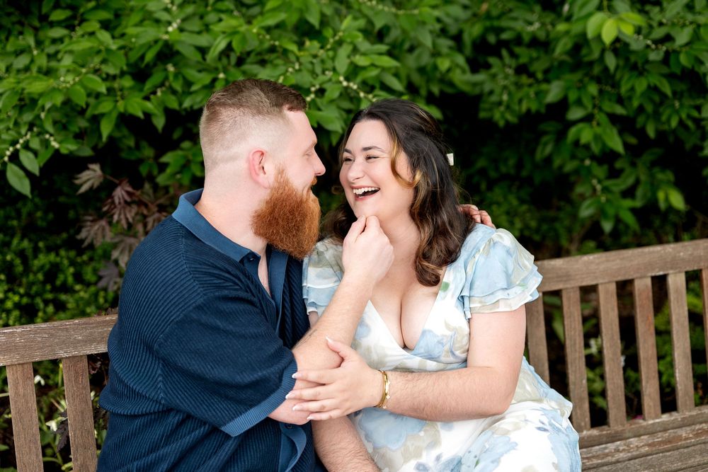 Couple laughing together on a wooden bench surrounded by lush green foliage during an outdoor engagement session.
