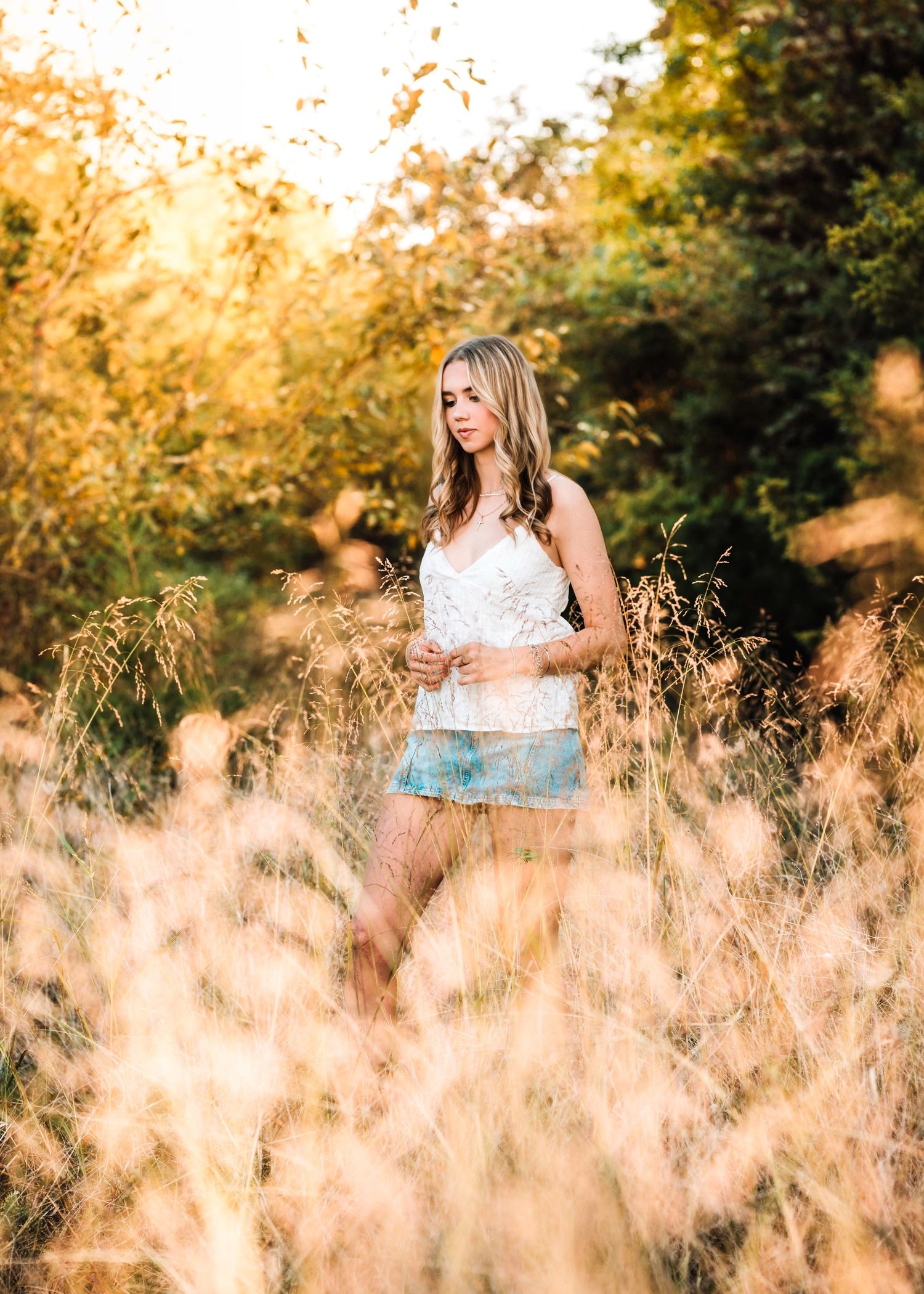 Young woman in white top and denim shorts standing in golden tall grass with warm sunlight filtering through trees.
