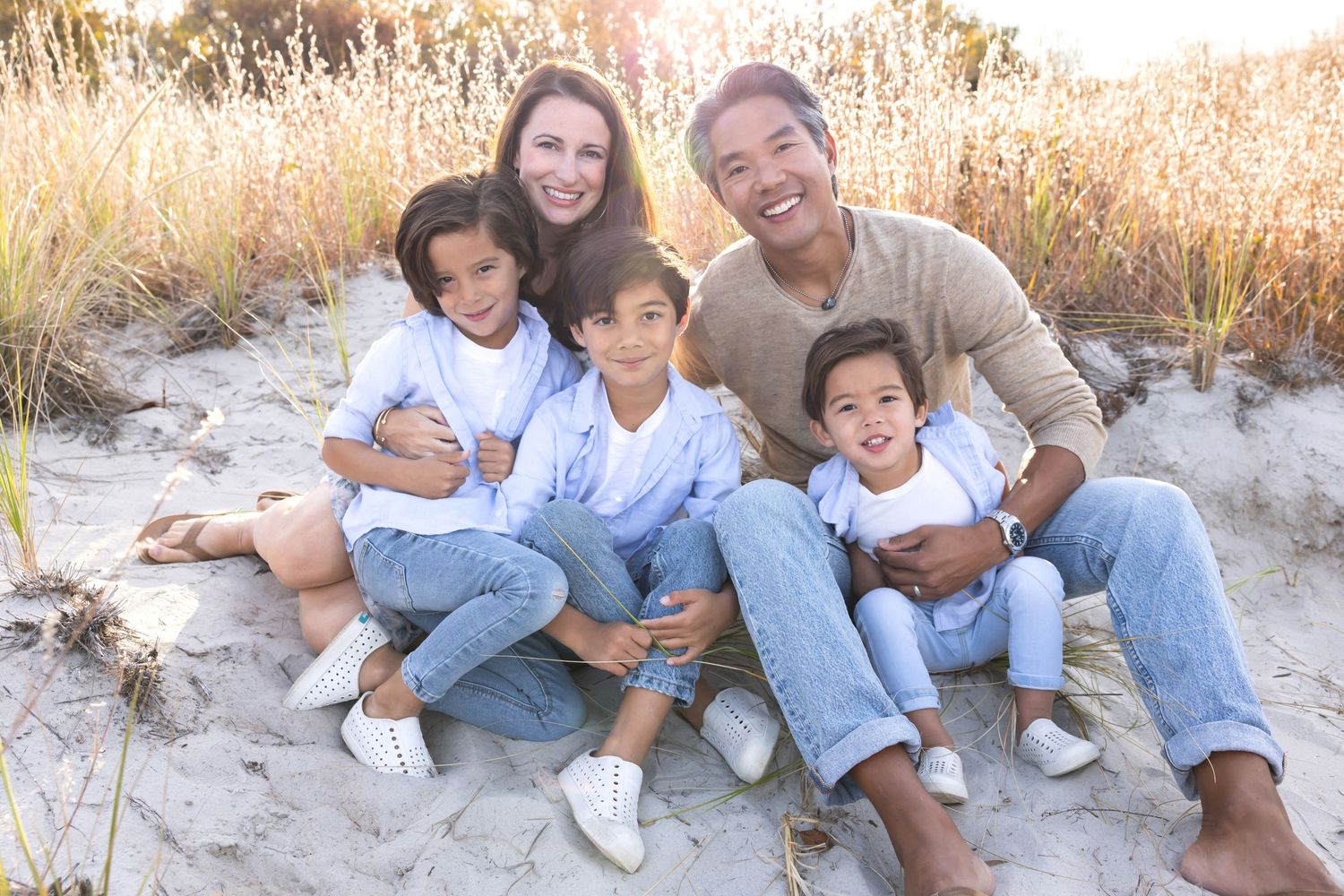 Happy multiracial family of five sitting together on sandy beach dunes during golden hour sunset.