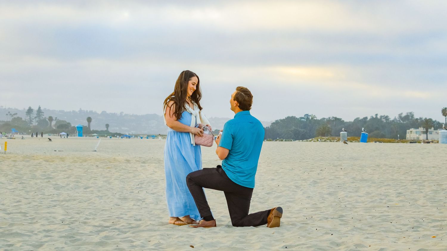 Man proposes on bended knee to woman in blue dress on sandy beach with cloudy sky backdrop.