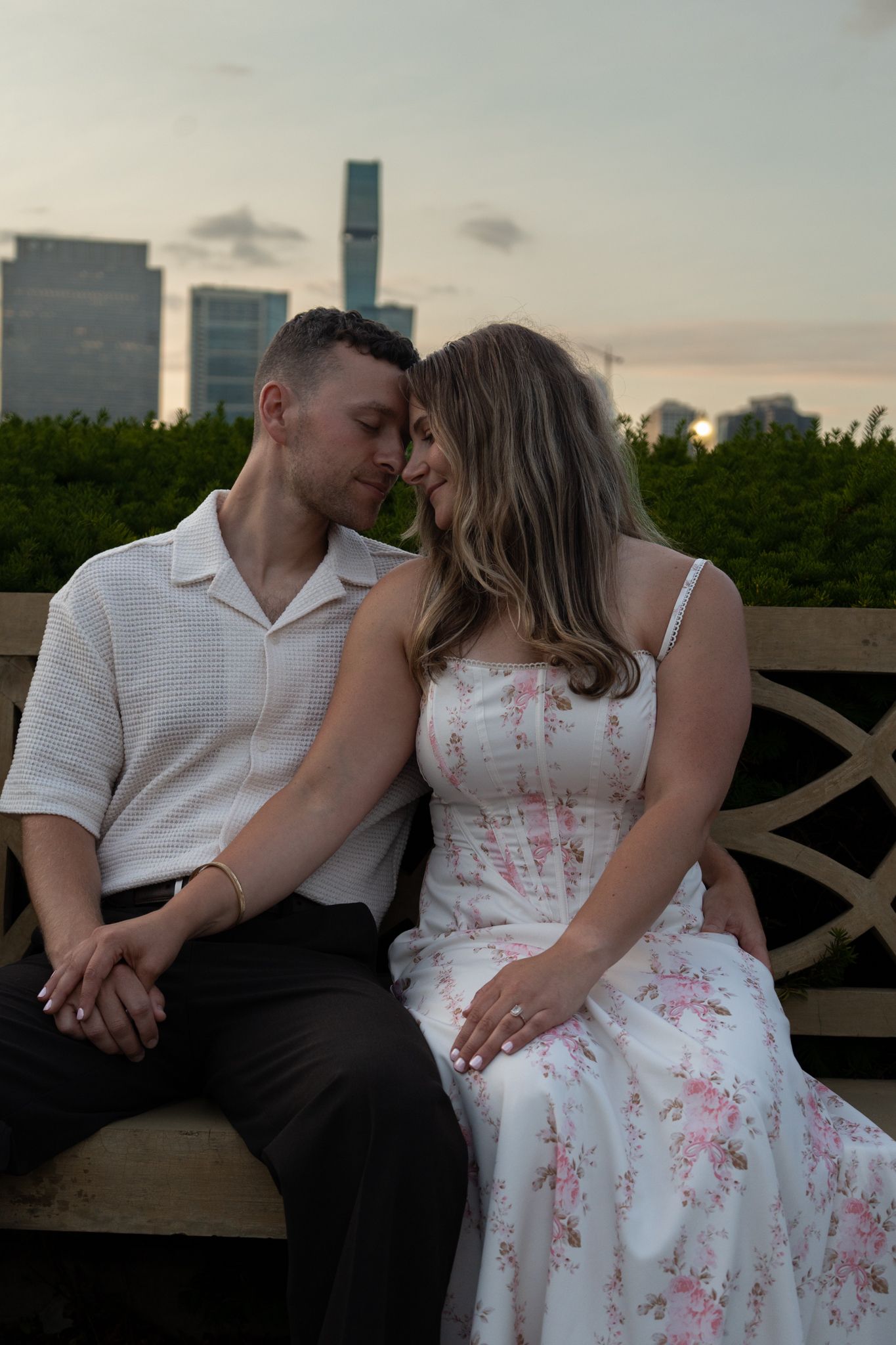 Couple sitting close on a wooden bench with city skyline at dusk, man in white shirt and woman in floral dress.