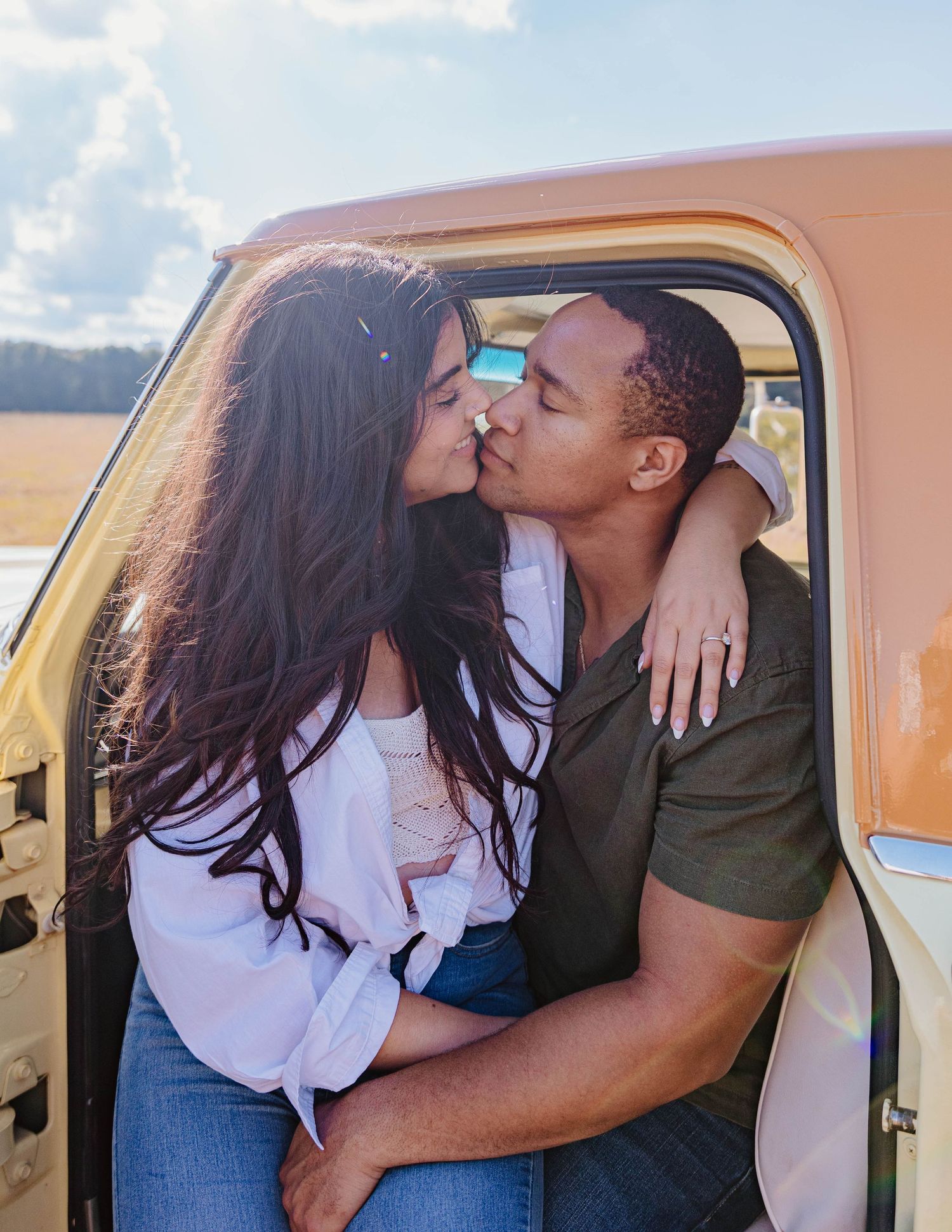 A couple shares a romantic kiss sitting in the open door of a vintage truck in a sunny rural field.