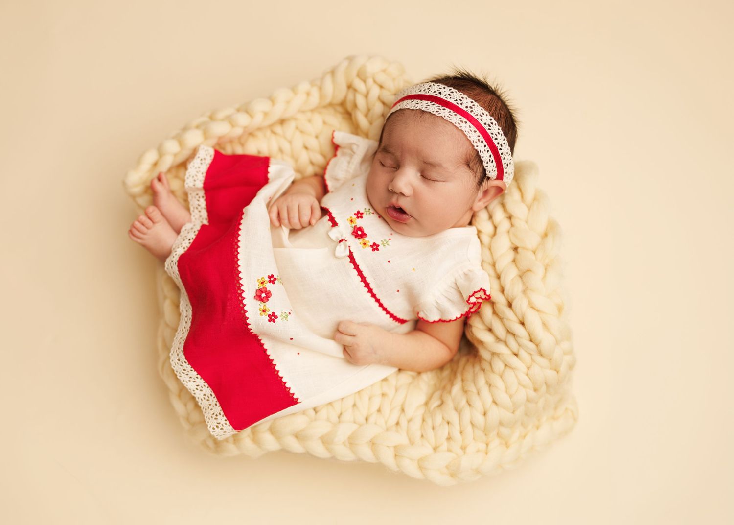 Sleeping newborn baby girl in white and red dress with matching headband nestled in chunky knit blanket.