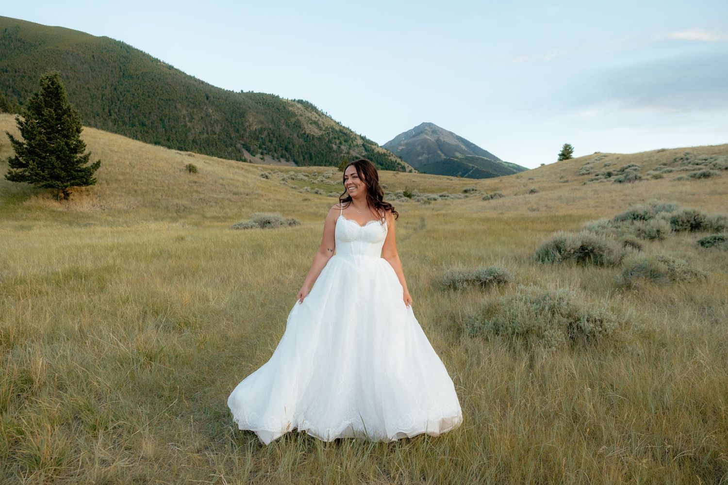 Bride in white ball gown wedding dress standing in scenic mountain meadow with golden grass and hills.