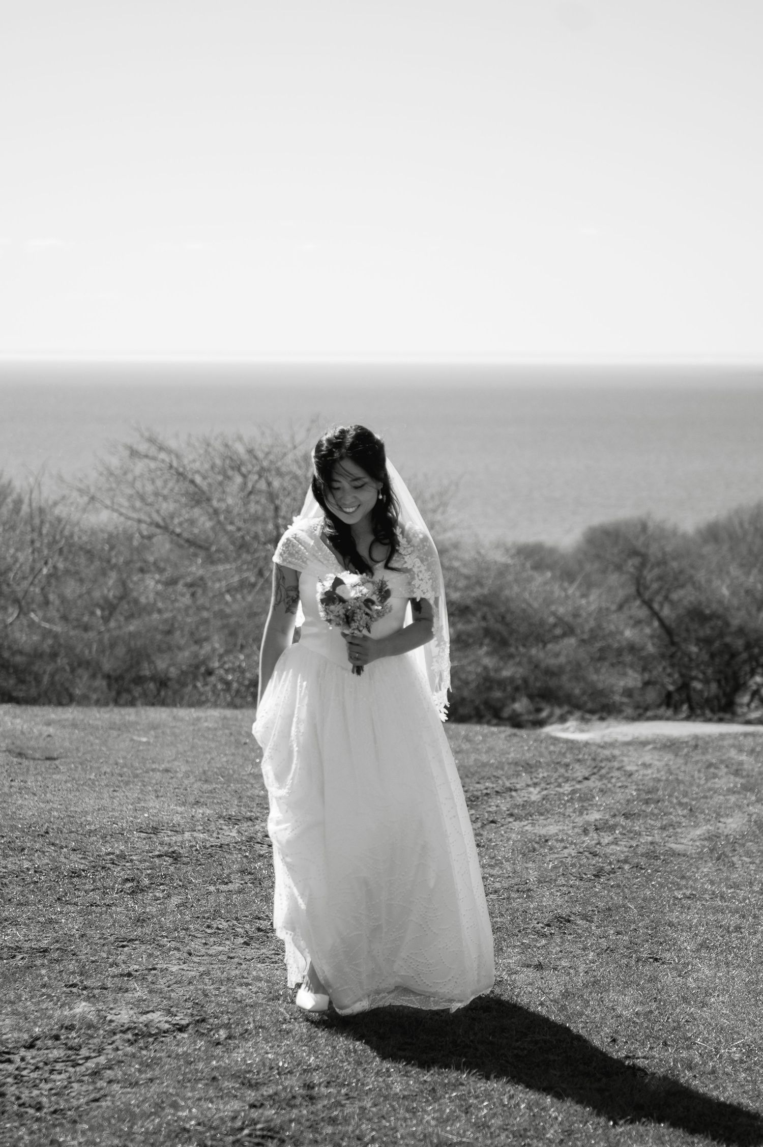 A bride in a white gown and veil holds a bouquet, smiling outdoors near the ocean in a black and white photo.
