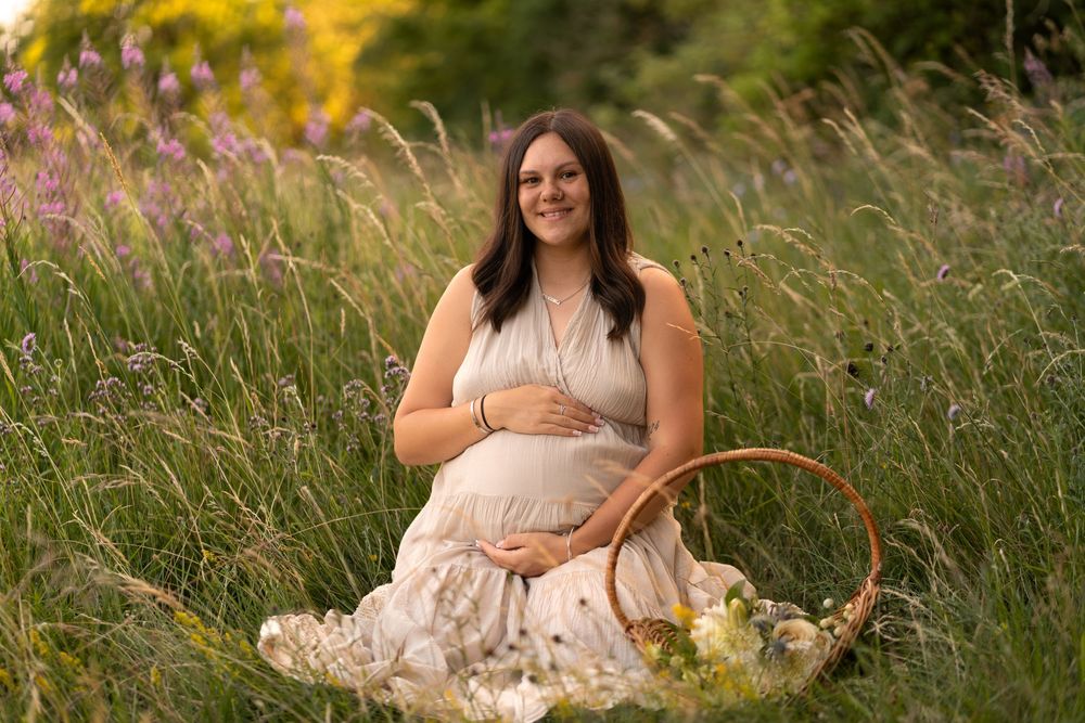 Smiling pregnant woman in beige dress sitting in wildflower field holding basket during golden hour maternity photo session.
