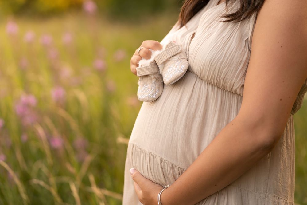 Close-up of pregnant woman in beige dress holding tiny baby shoes over her baby bump in a wildflower meadow.