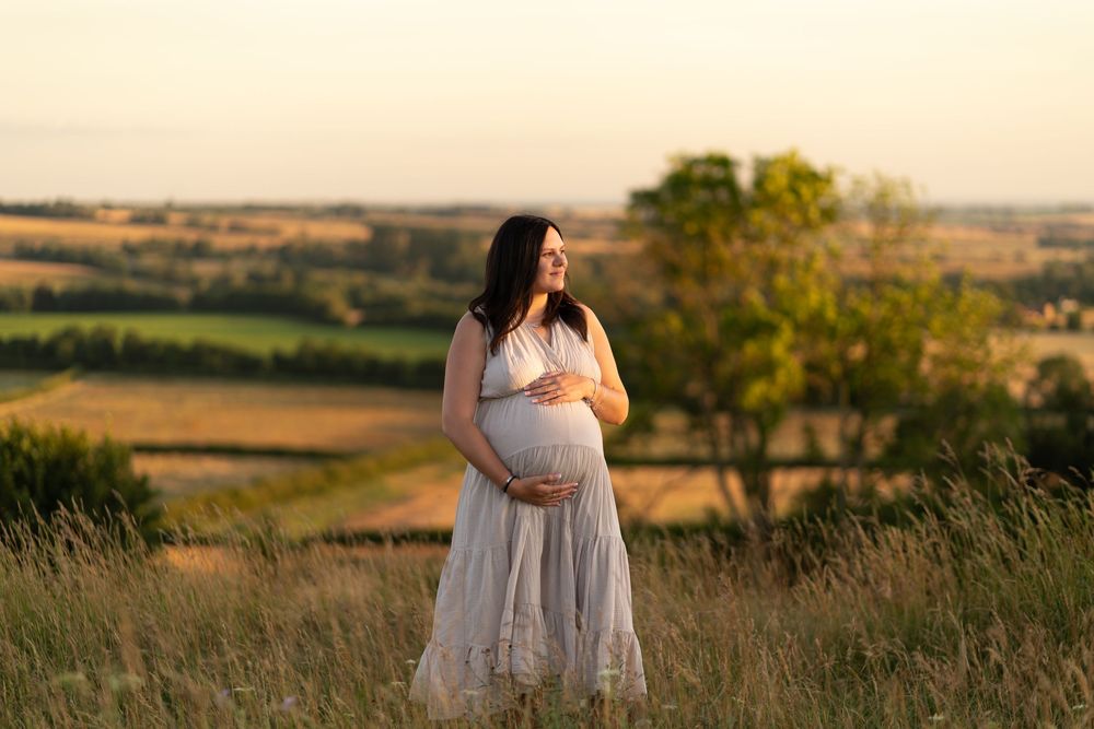 Pregnant woman in white dress holding baby bump at golden hour in scenic countryside field.