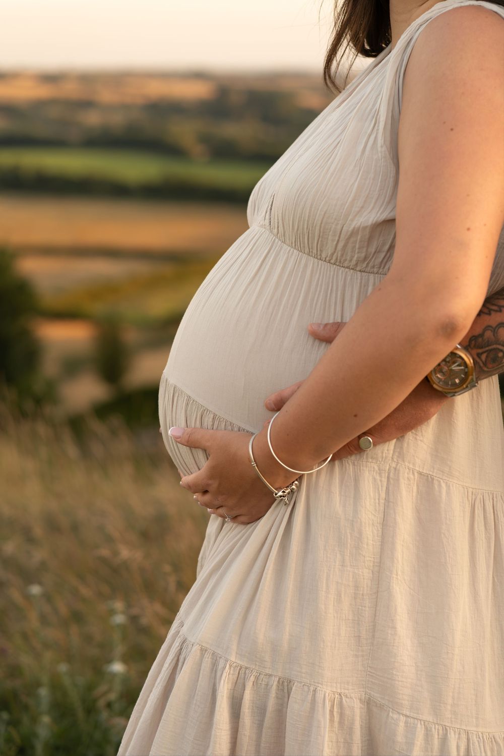 Pregnant woman in beige dress cradling baby bump outdoors at golden hour with scenic countryside backdrop.