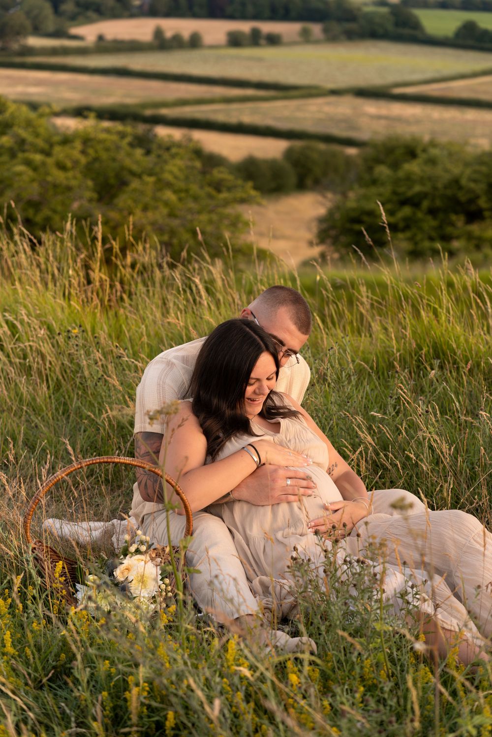 Couple sitting in golden hour meadow, man embracing pregnant woman with picnic basket and wildflowers, rolling countryside behind them.