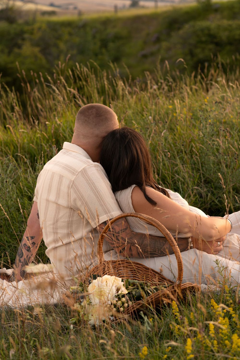 Couple sitting together in a meadow at sunset with a wicker picnic basket and white flowers.