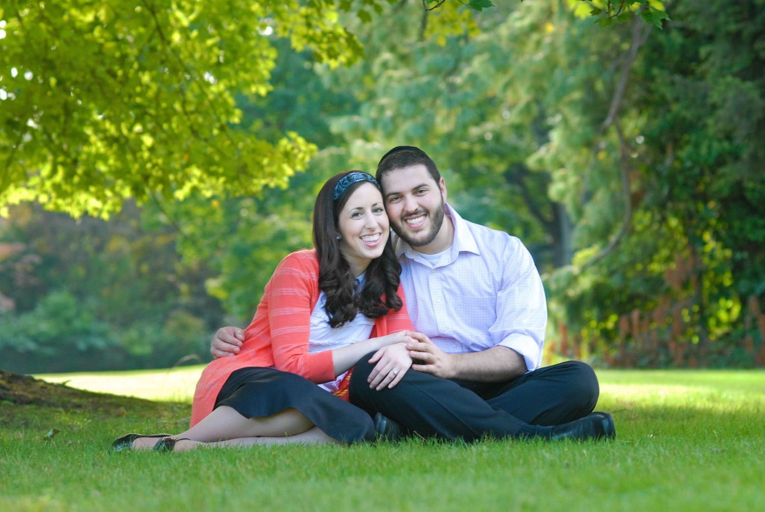 Happy couple sitting on green grass in a park, surrounded by lush trees, smiling and embracing outdoors.