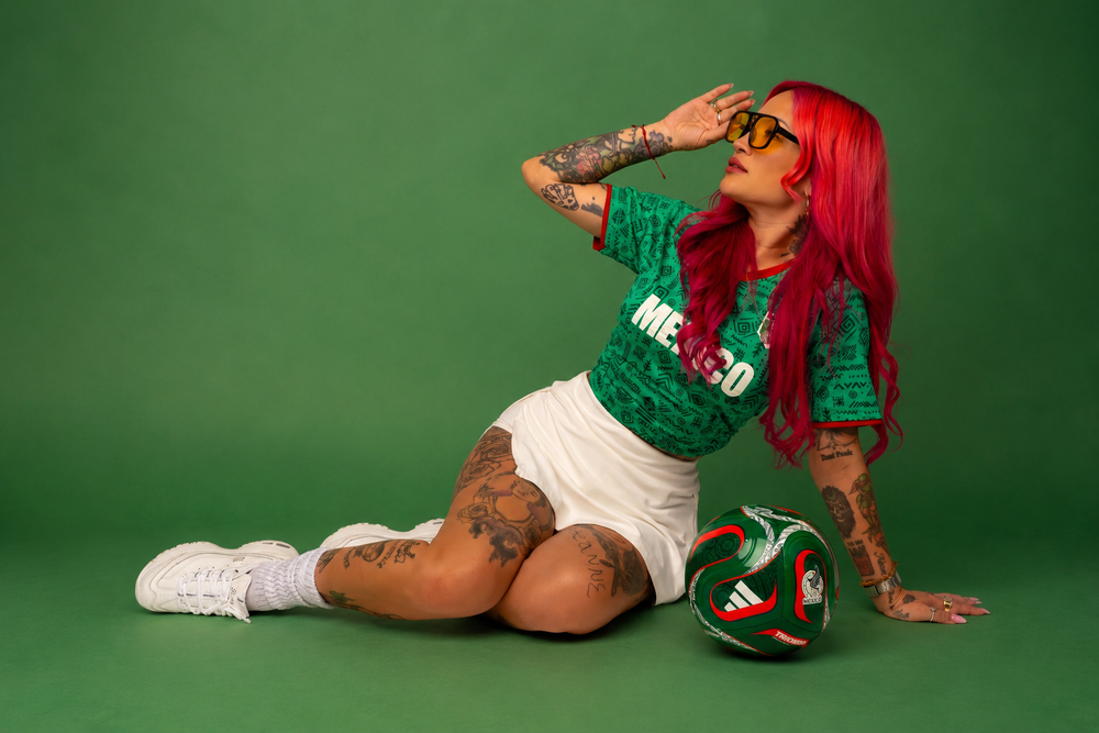Woman with red hair in Mexico soccer jersey sits with a soccer ball on a green background.
