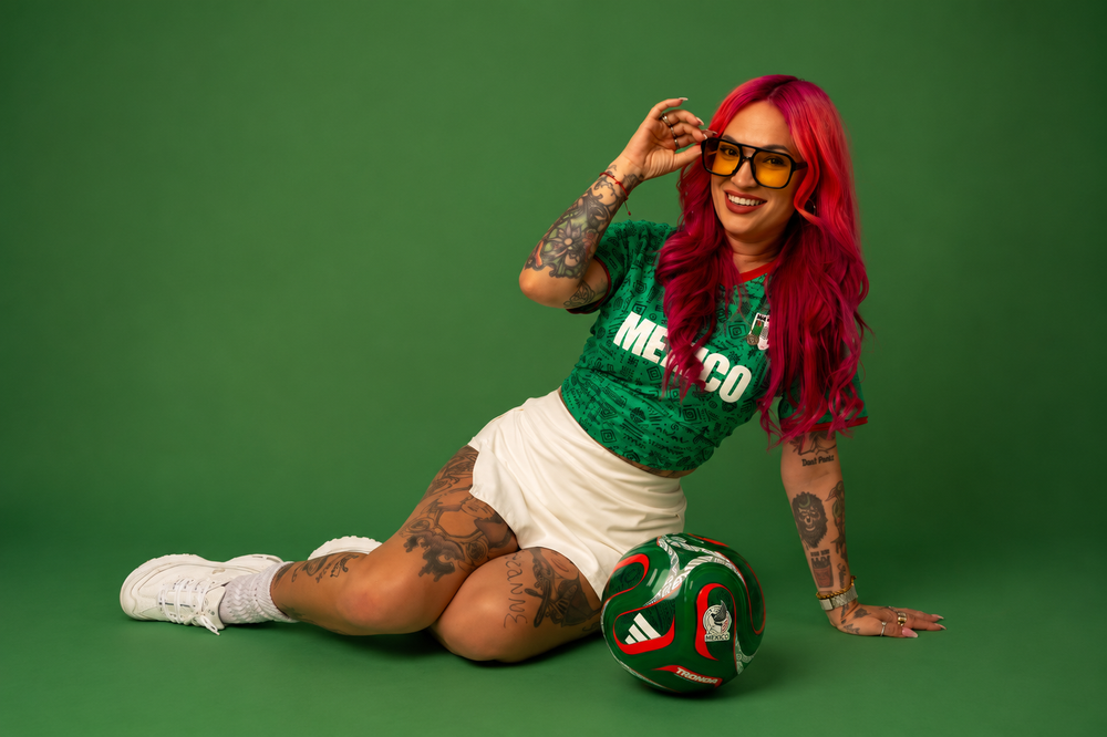 Woman with red hair in Mexico soccer jersey and white skirt sits with Adidas soccer ball on green background.