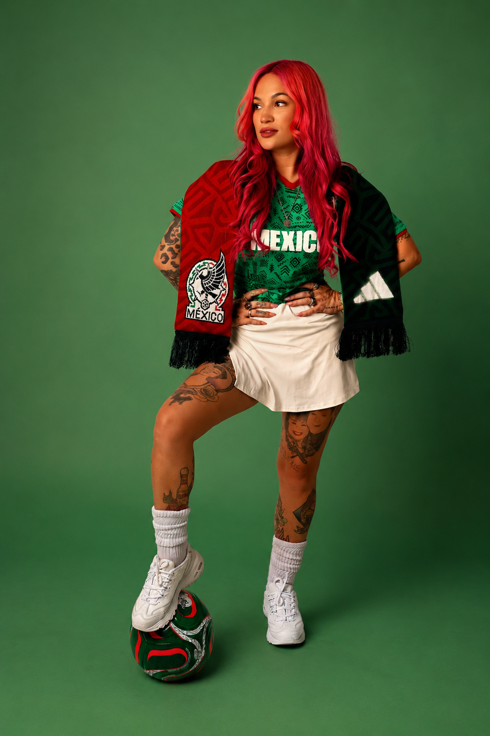 Red-haired woman in Mexico soccer jersey holds scarf while posing with Adidas ball on green background.