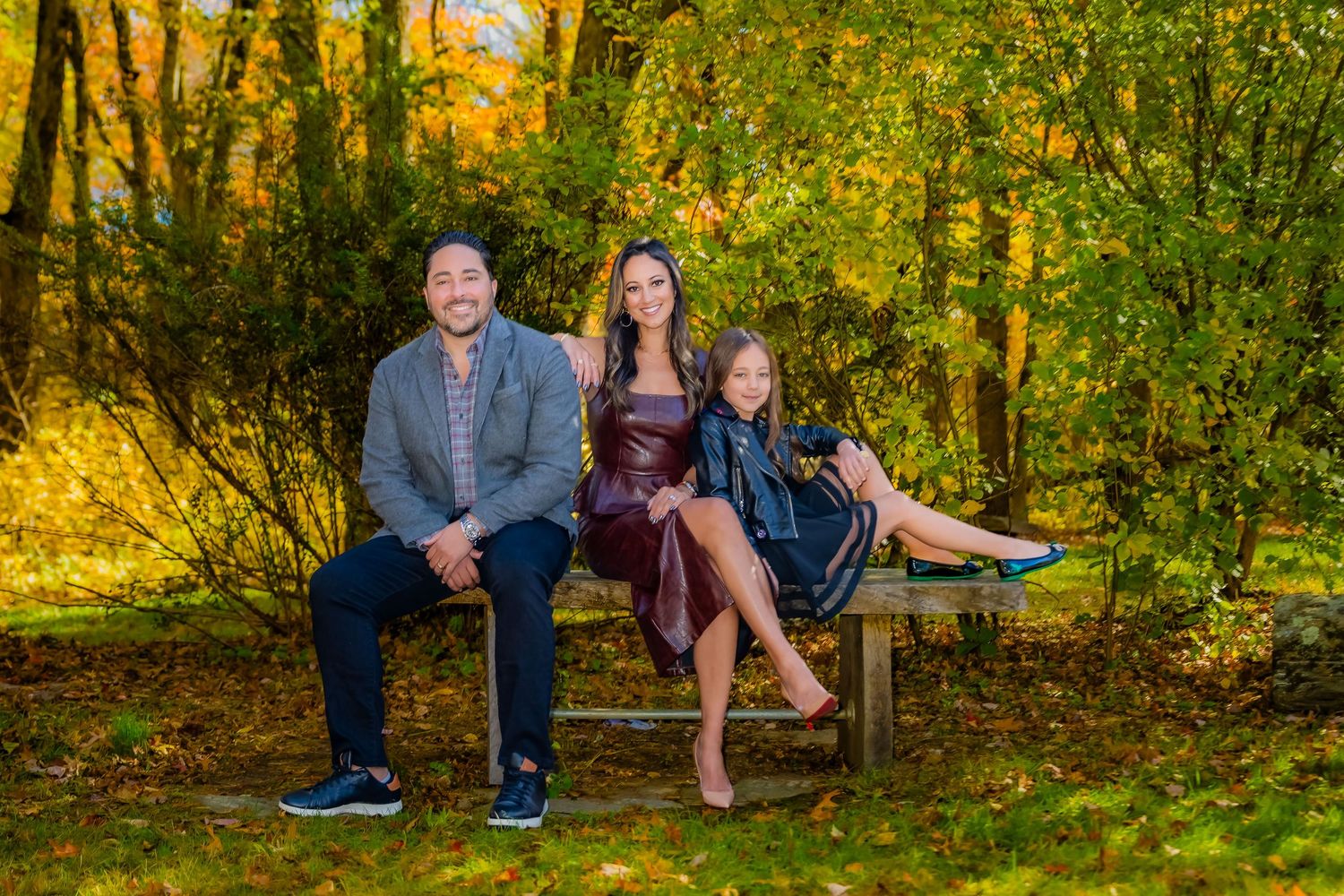Family of three smiling on a bench surrounded by vibrant autumn foliage in a lush green park setting.
