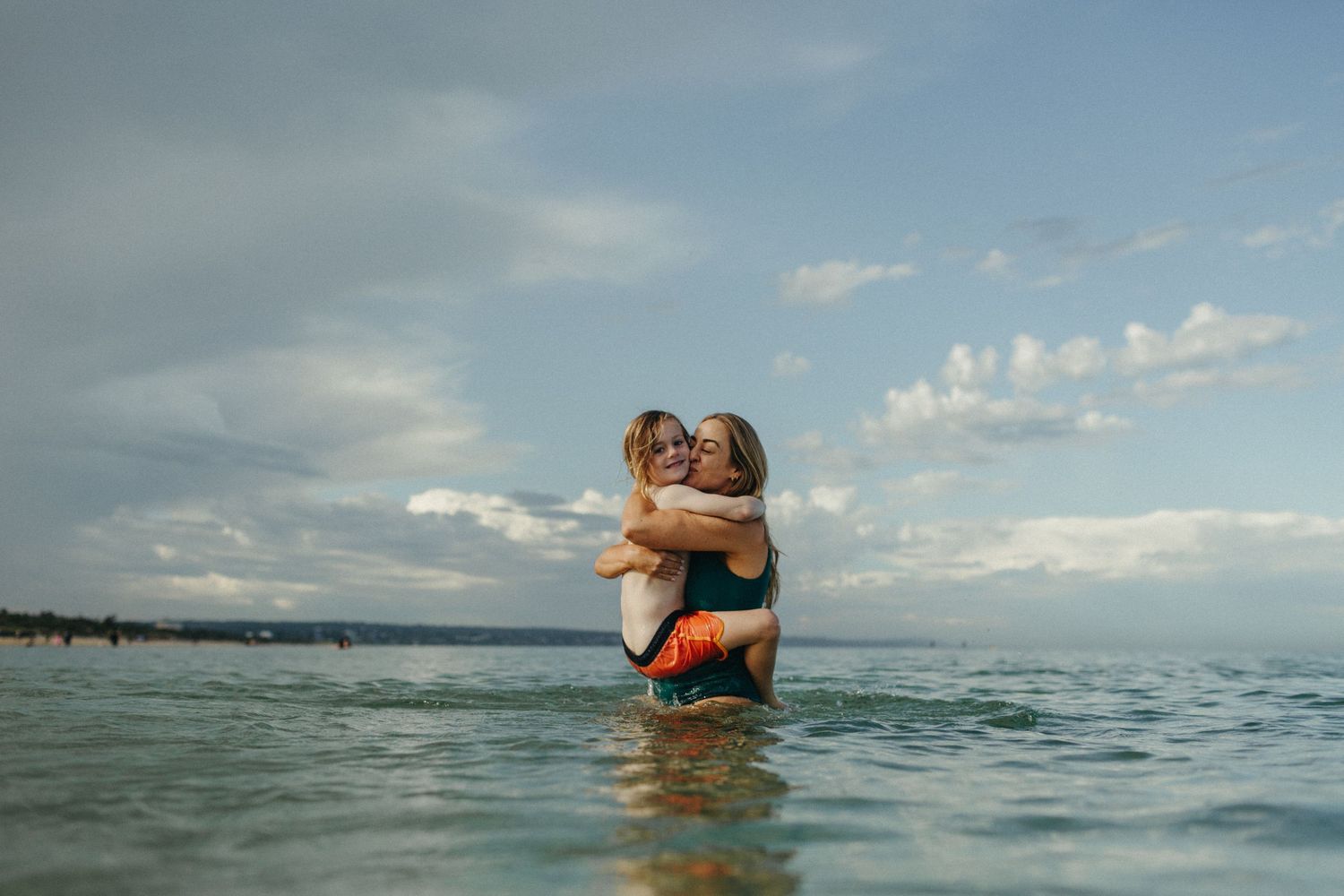Black and white photo of a woman hugging a child in shallow ocean water near a sandy beach.