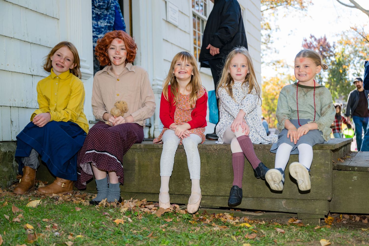 Five young girls in colorful costumes sit together on outdoor steps during an autumn day.