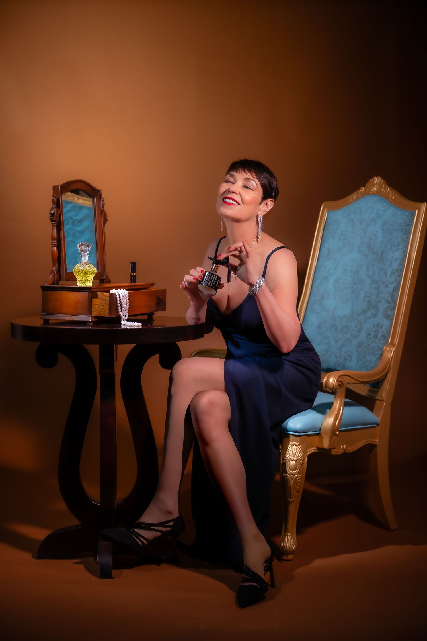 Elegant woman in blue dress sits by vintage vanity table with perfume bottles and ornate gold chair in warm studio setting.