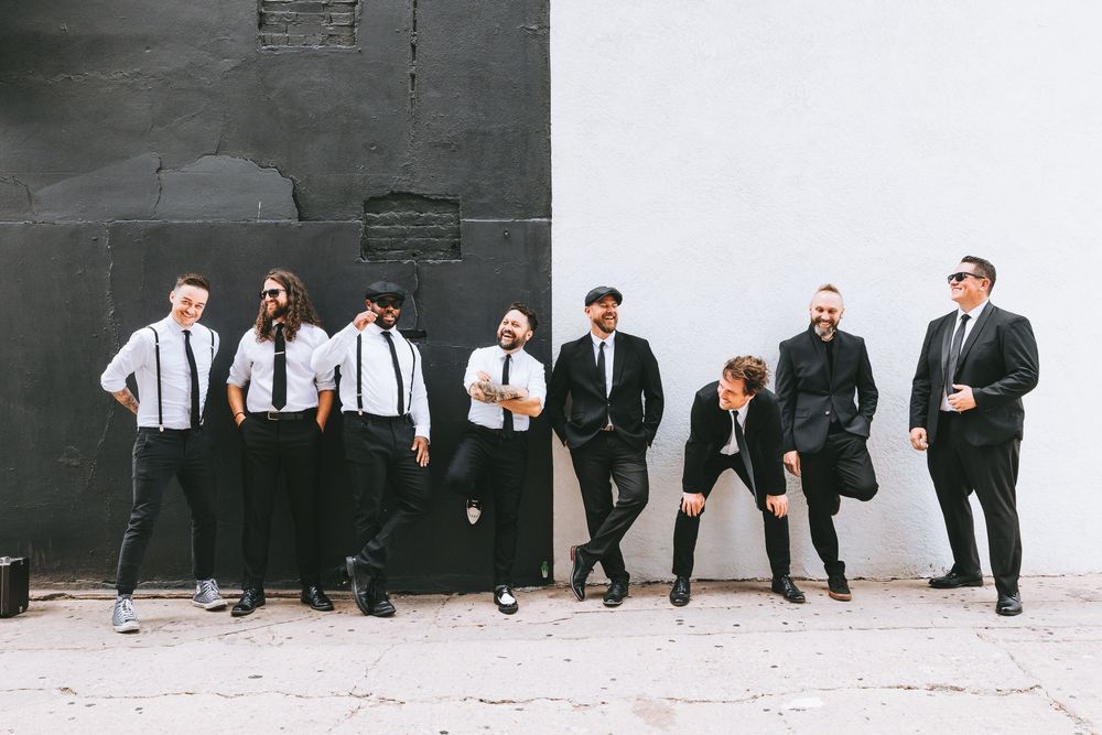 Group of men in black suits and suspenders posing against a black and white wall outdoors.