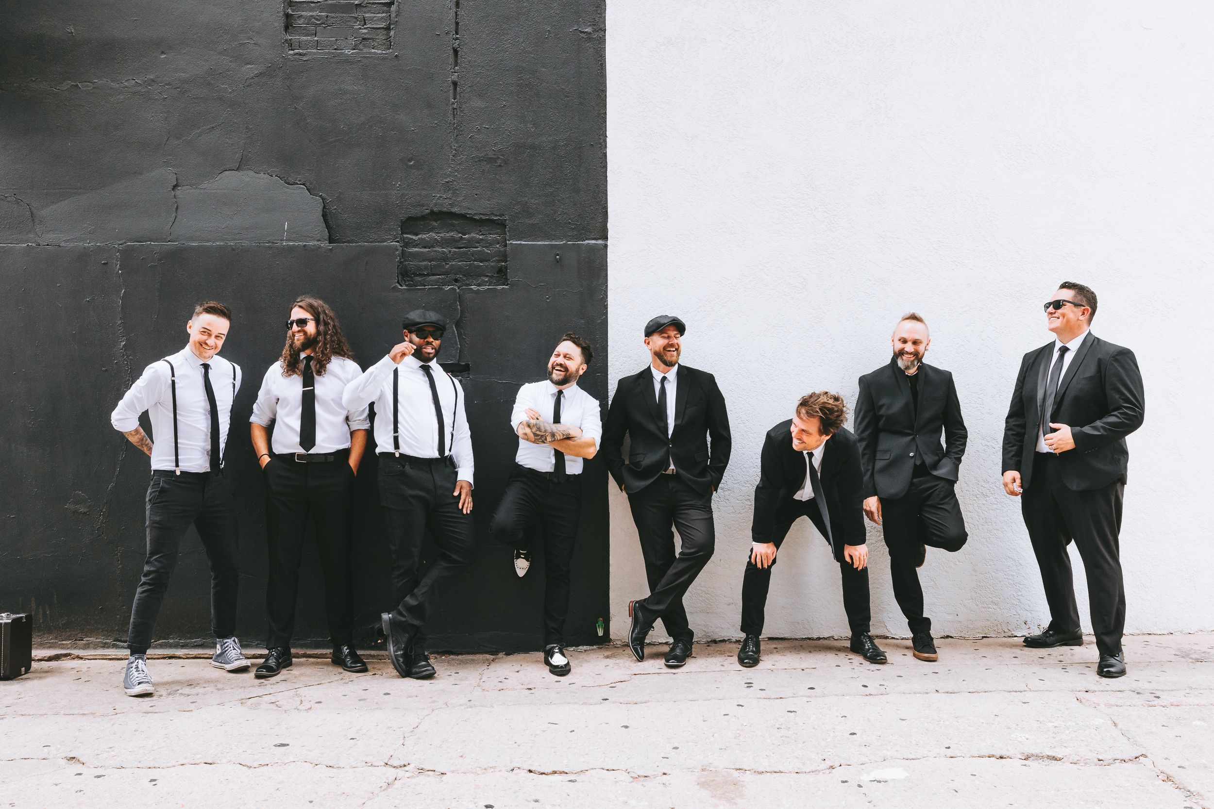 Group of men in black suits and suspenders posing against a black and white wall outdoors.