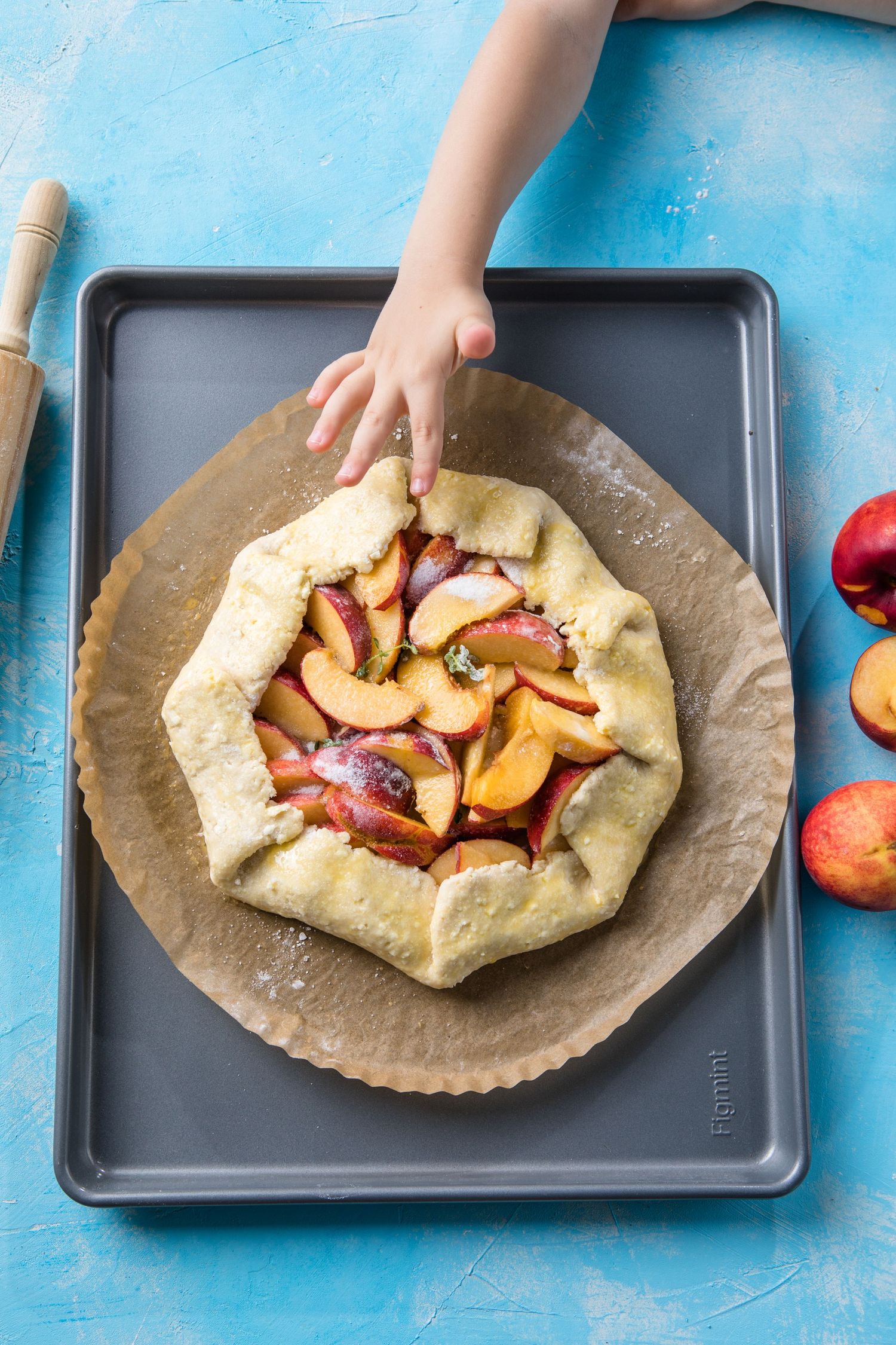 Child's hand touching a rustic peach galette on parchment paper on a baking sheet, with fresh peaches nearby.