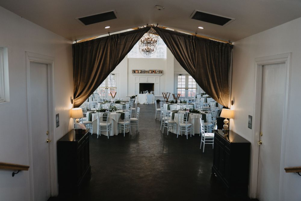 Elegant wedding reception hall with draped curtains, round tables, white chairs, and a chandelier in a spacious venue.