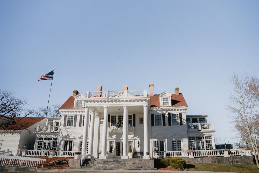 Grand white colonial mansion with large columns, American flag, and red-roofed dormers under a clear blue sky.