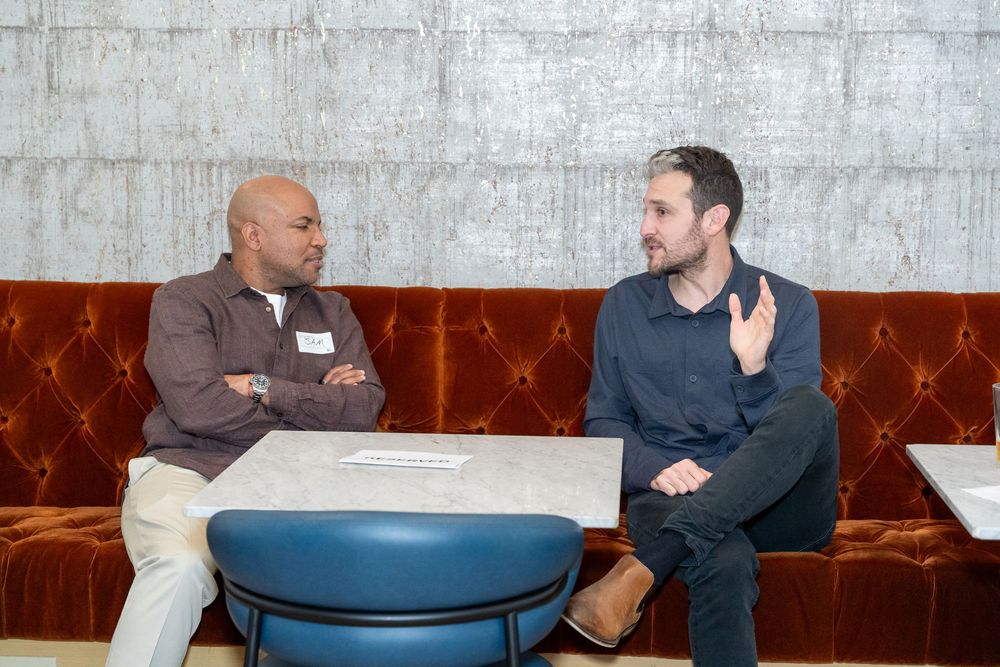 Two men having a conversation on a red velvet sofa at a modern lounge, one wearing a name tag.