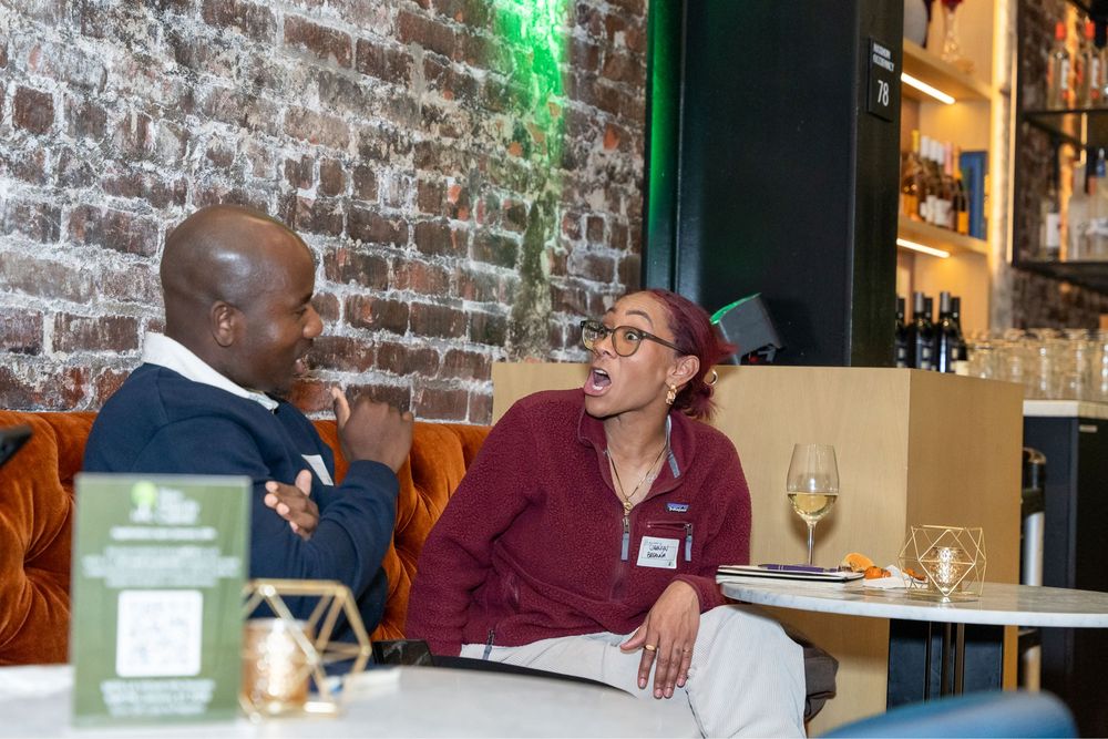 Two people engaged in lively conversation at a restaurant bar with exposed brick wall and wine glasses.