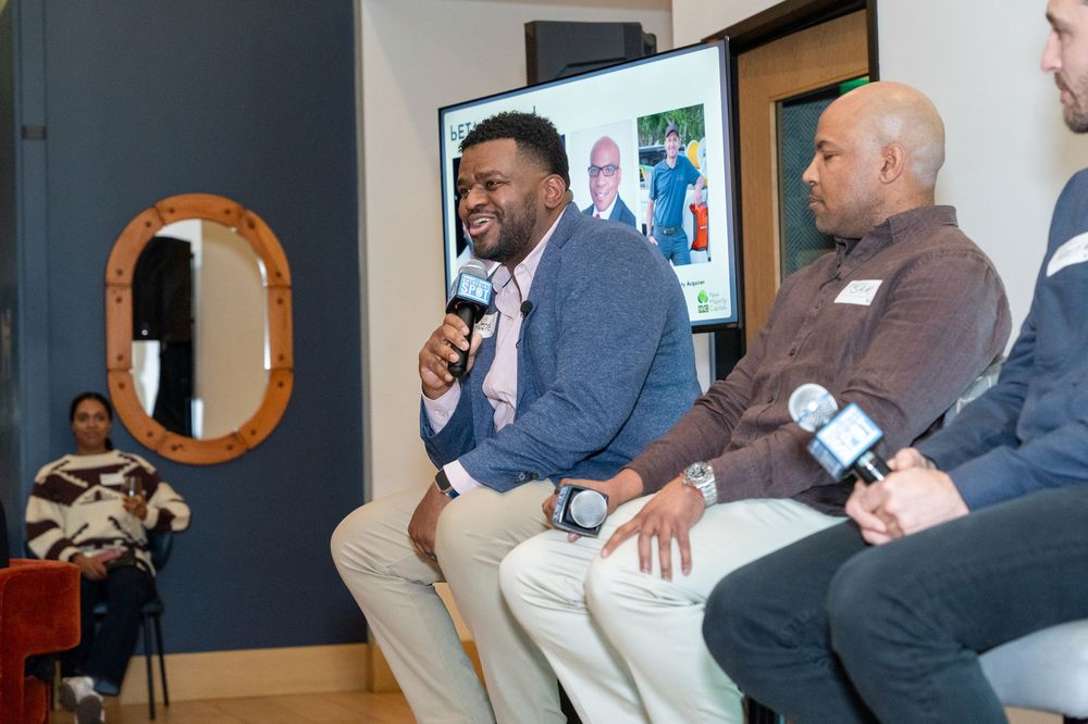 Three men seated at a panel discussion, one speaking into a microphone, with an audience member visible in the background.