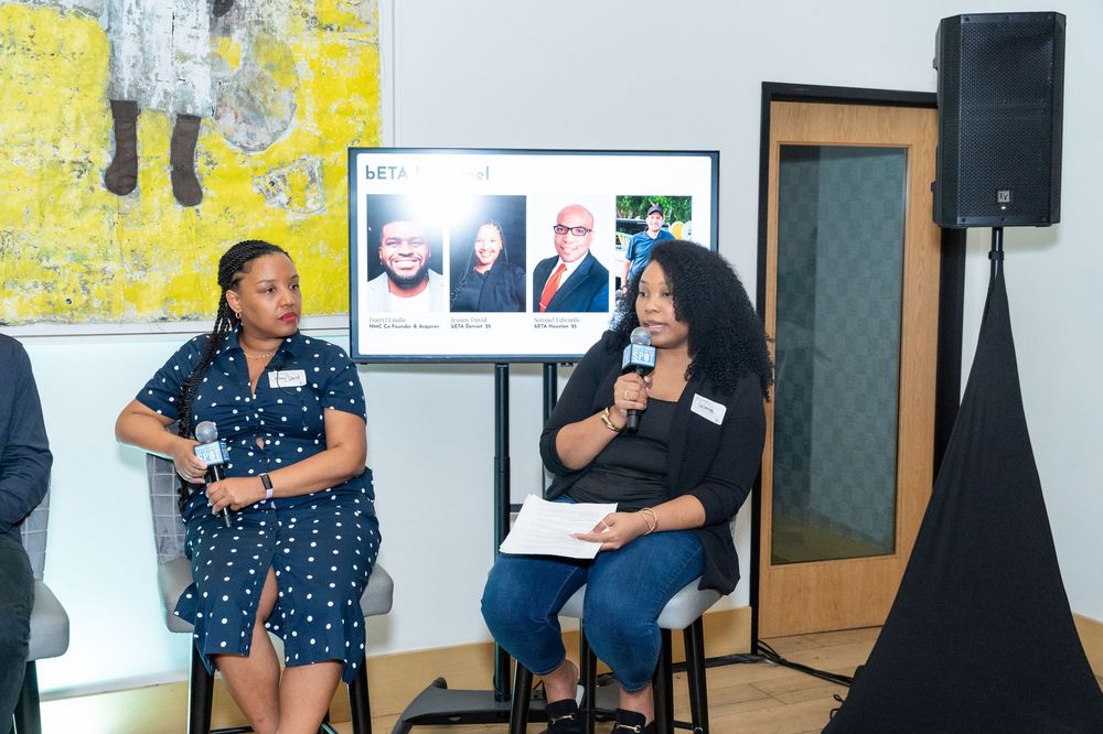 Two women sit on stools at an indoor panel event, one holding a microphone, with a presentation screen visible behind them.