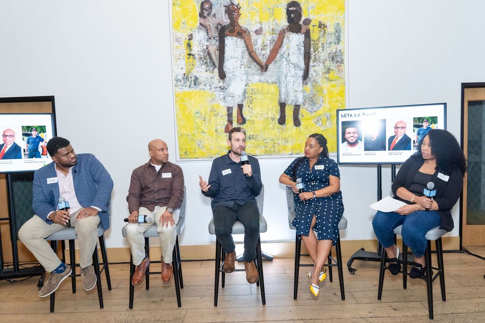 Five panelists seated on stools in discussion at an indoor event with colorful artwork displayed on the wall behind them.