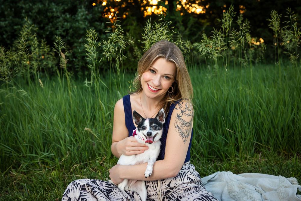 Smiling tattooed woman sitting on blanket with small dog in sunny field.